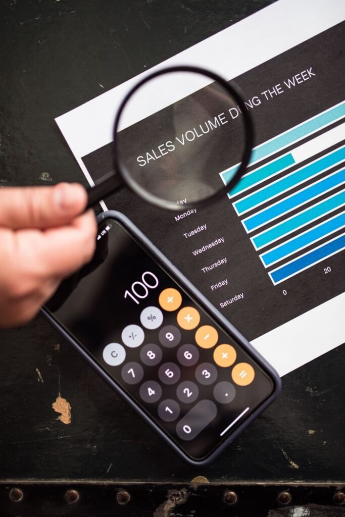 Magnifying glass focuses on sales graph and smartphone calculator, depicting data analysis.